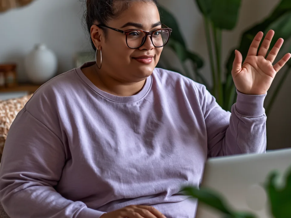 A woman in a lavender sweatshirt smiling and waving hello during a virtual session