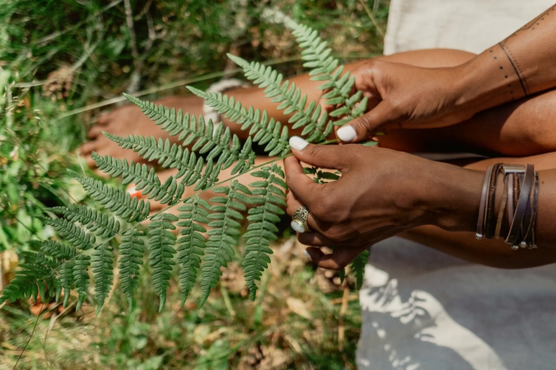 Hands holding a fern frond, with stacked leather bracelets and cream linen
