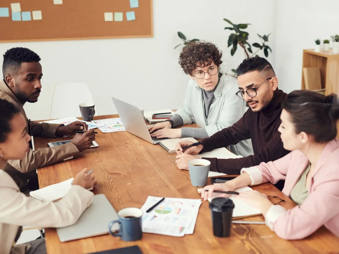 A diverse team of colleagues in discussion around a wooden conference table with laptops, notes, and coffee
